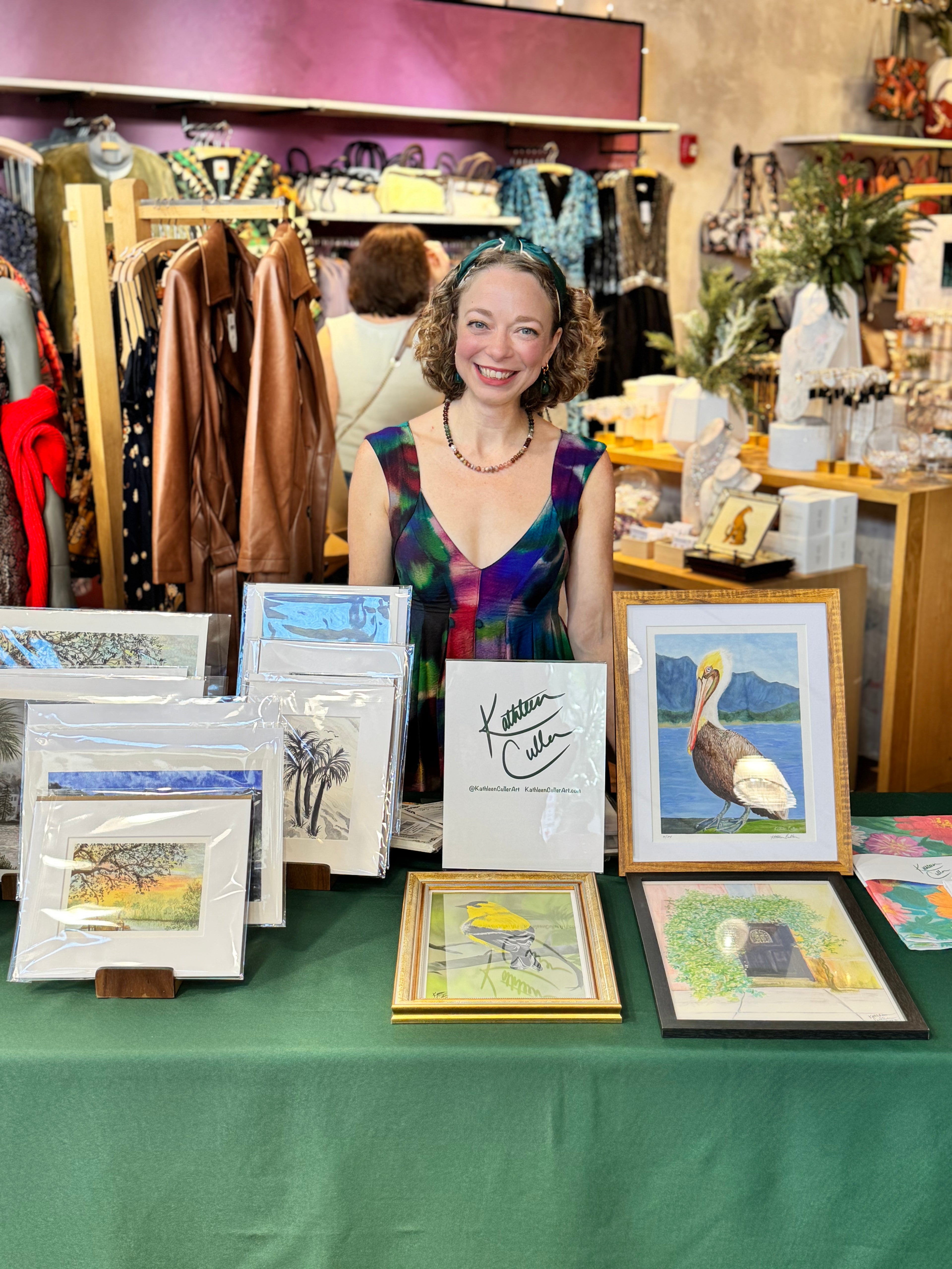Artist Kathleen Culler standing behind a table displaying various framed artworks in a store setting.